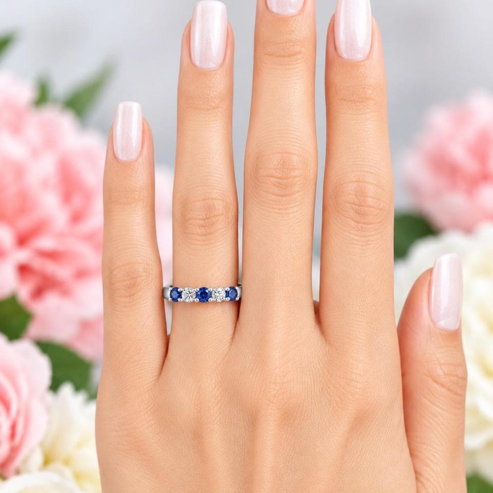 A woman's hand wearing a sterling silver 5-stone band featuring alternating round lab blue sapphires and moissanites. The band has a classic design with the gemstones set in a row with three sapphires and two moissanites. Model image for size and styling reference — not a photo of the actual ring.
