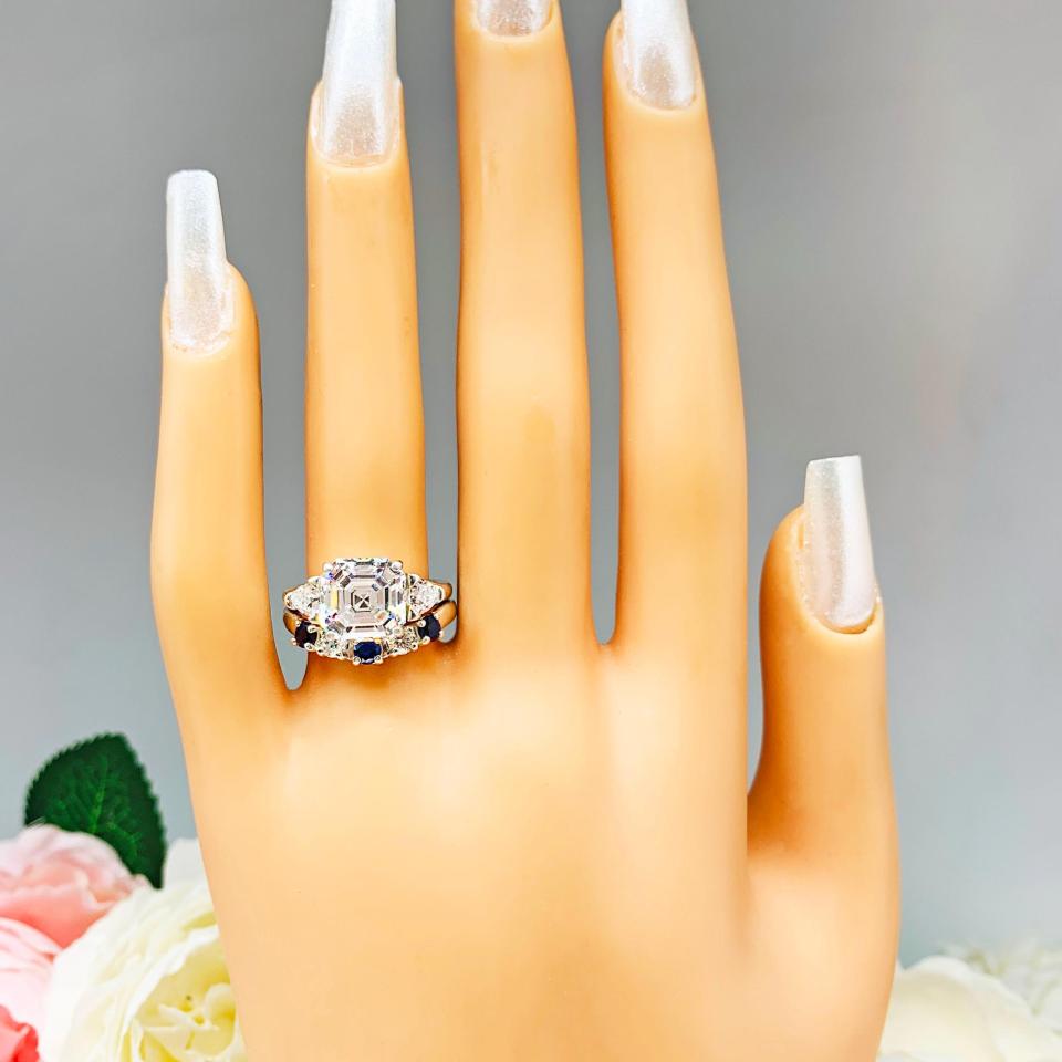 Mannequin hand with long white shimmery nails, displayed against a soft white-gray background with pink and white floral accents. The hand is wearing a sterling silver 5-stone band featuring alternating round lab blue sapphires and moissanites. The band has a classic design with the gemstones set in a row with three sapphires and two moissanites. It is featured here with an asscher-cut cz with trillion sides three stone ring (not included).