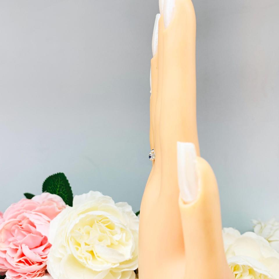Mannequin hand with long white shimmery nails, displayed against a soft white-gray background with pink and white floral accents. The hand is wearing a sterling silver curved 5-stone band featuring alternating round lab blue sapphires and moissanites. The band has a classic design with the gemstones set in a row with three sapphires and two moissanites.