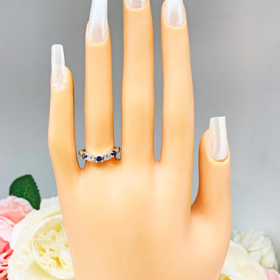 Mannequin hand with long white shimmery nails, displayed against a soft white-gray background with pink and white floral accents. The hand is wearing a sterling silver curved 5-stone band featuring alternating round lab blue sapphires and moissanites. The band has a classic design with the gemstones set in a row with three sapphires and two moissanites.