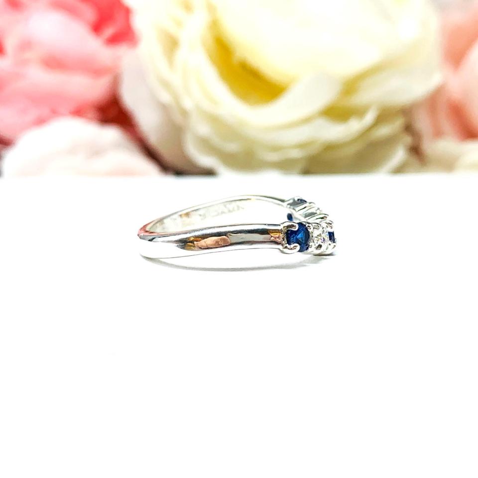 A sterling silver curved 5-stone band featuring alternating round lab blue sapphires and moissanites. The band has a classic design with the gemstones set in a row with three sapphires and two moissanites. Displayed on a white surface with a pastel floral background.
