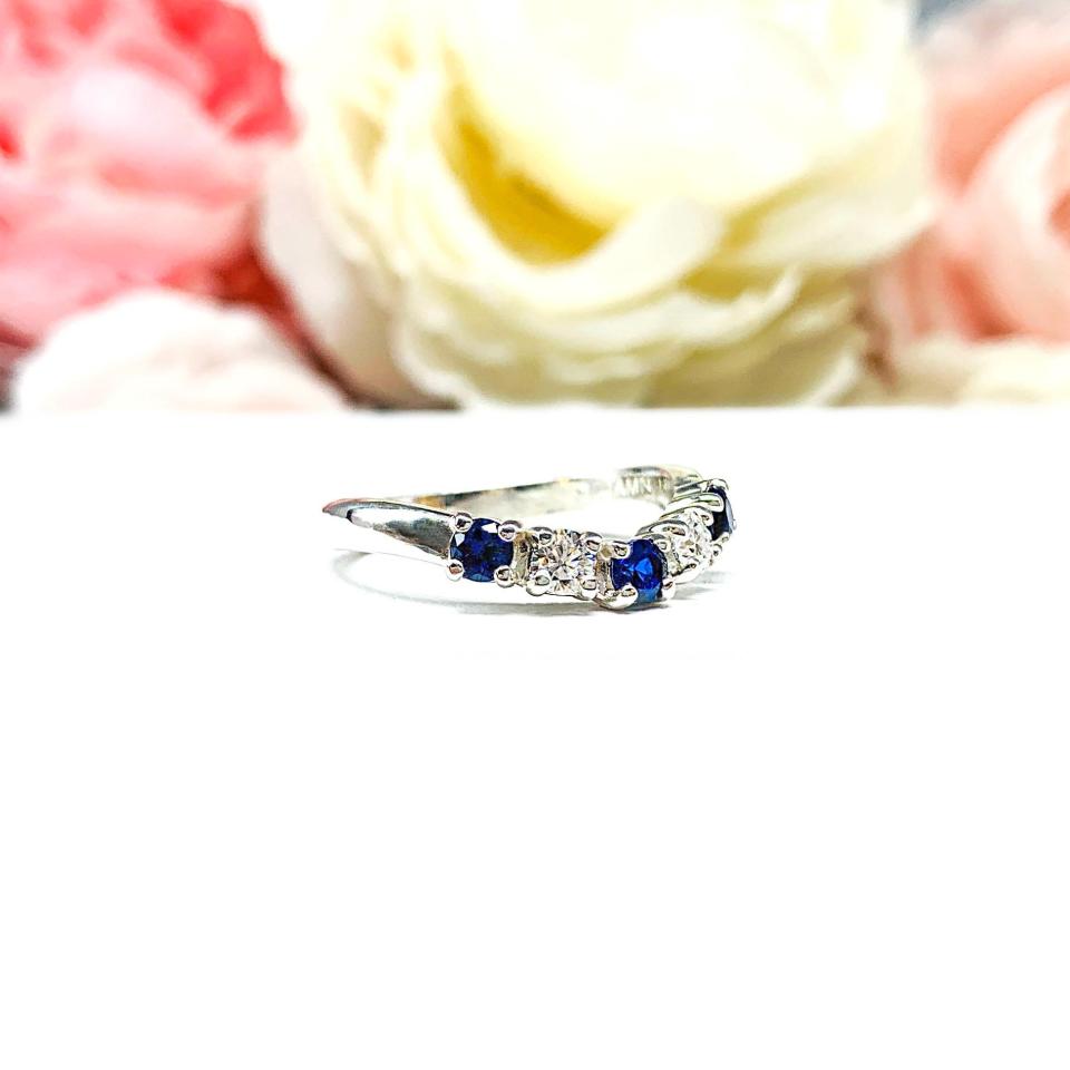 A sterling silver curved 5-stone band featuring alternating round lab blue sapphires and moissanites. The band has a classic design with the gemstones set in a row with three sapphires and two moissanites. Displayed on a white surface with a pastel floral background.