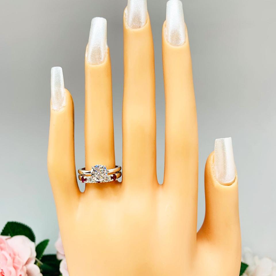 Mannequin hand with long white shimmery nails, displayed against a soft white-gray background with pink and white floral accents. The hand is wearing a sterling silver 5-stone band featuring alternating round lab rubies and moissanites. The band has a classic design with the gemstones set in a row with three rubies and two moissanites. Shown with a round moissanite solitaire ring (not included).