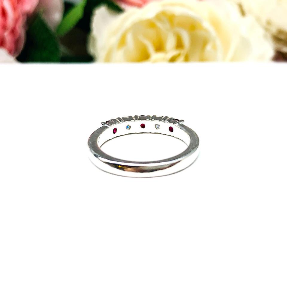 A sterling silver 5-stone band featuring alternating round lab rubies and moissanites. The band has a classic design with the gemstones set in a row with three rubies and two moissanites. Displayed on a white surface with a pastel floral background.