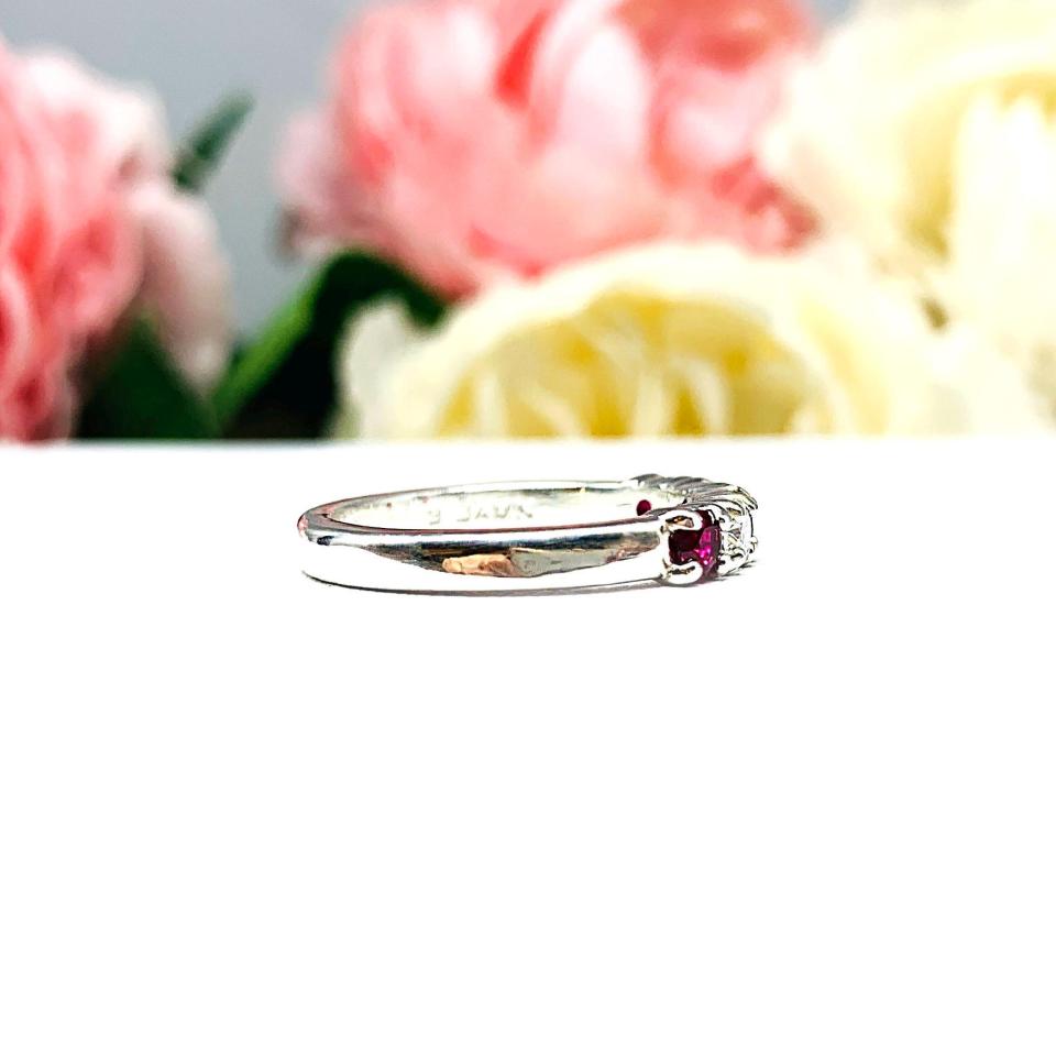 A sterling silver 5-stone band featuring alternating round lab rubies and moissanites. The band has a classic design with the gemstones set in a row with three rubies and two moissanites. Displayed on a white surface with a pastel floral background.