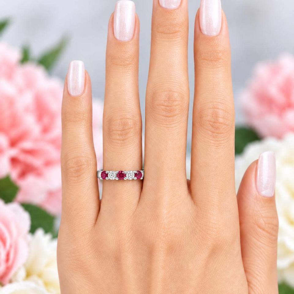 A woman's hand wearing a sterling silver 5-stone band featuring alternating round lab rubies and moissanites. The band has a classic design with the gemstones set in a row with three rubies and two moissanites. Model image for size and styling reference — not a photo of the actual ring.