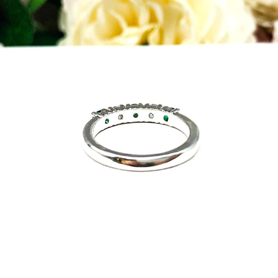 Sterling silver five-stone wedding band with three 3mm round lab-created emeralds and two 3mm round DEF moissanites, shown against a soft floral background.