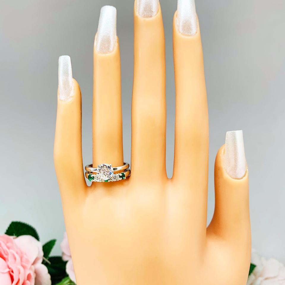 Mannequin hand with long white shimmery nails, displayed against a soft white-gray background with pink and white floral accents. The hand is wearing a sterling silver  five-stone wedding band with three 3mm round lab-created emeralds and two 3mm round DEF moissanites. Shown with a round moissanite solitaire ring (not included).