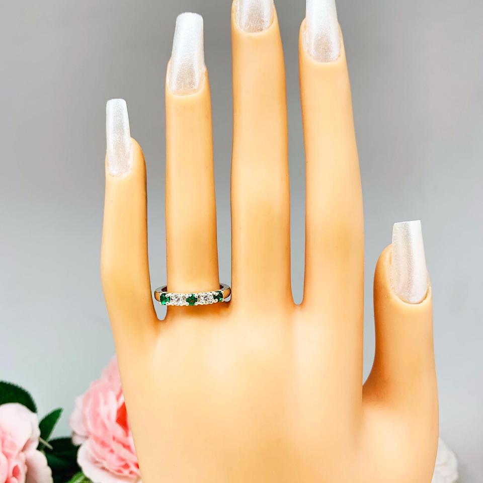 Mannequin hand with long white shimmery nails, displayed against a soft white-gray background with pink and white floral accents. The hand is wearing a sterling silver  five-stone wedding band with three 3mm round lab-created emeralds and two 3mm round DEF moissanites.