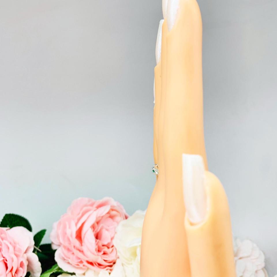 Mannequin hand with long white shimmery nails, displayed against a soft white-gray background with pink and white floral accents. The hand is wearing a sterling silver  five-stone wedding band with three 3mm round lab-created emeralds and two 3mm round DEF moissanites.