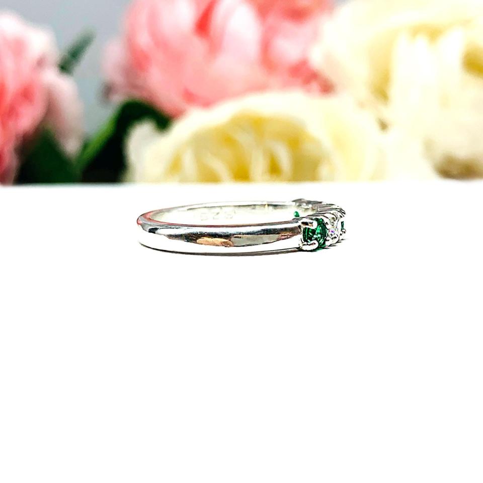 Sterling silver five-stone wedding band with three 3mm round lab-created emeralds and two 3mm round DEF moissanites, shown against a soft floral background.