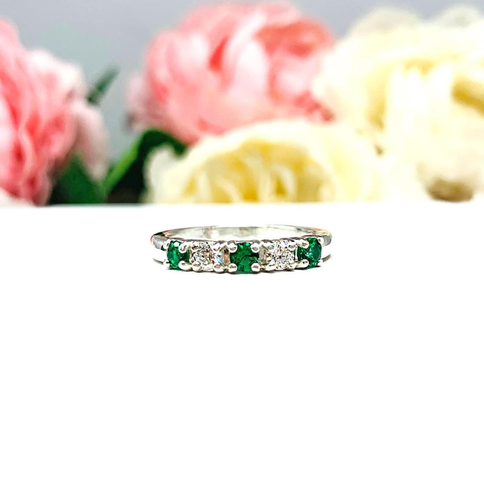 Sterling silver five-stone wedding band with three 3mm round lab-created emeralds and two 3mm round DEF moissanites, shown against a soft floral background.