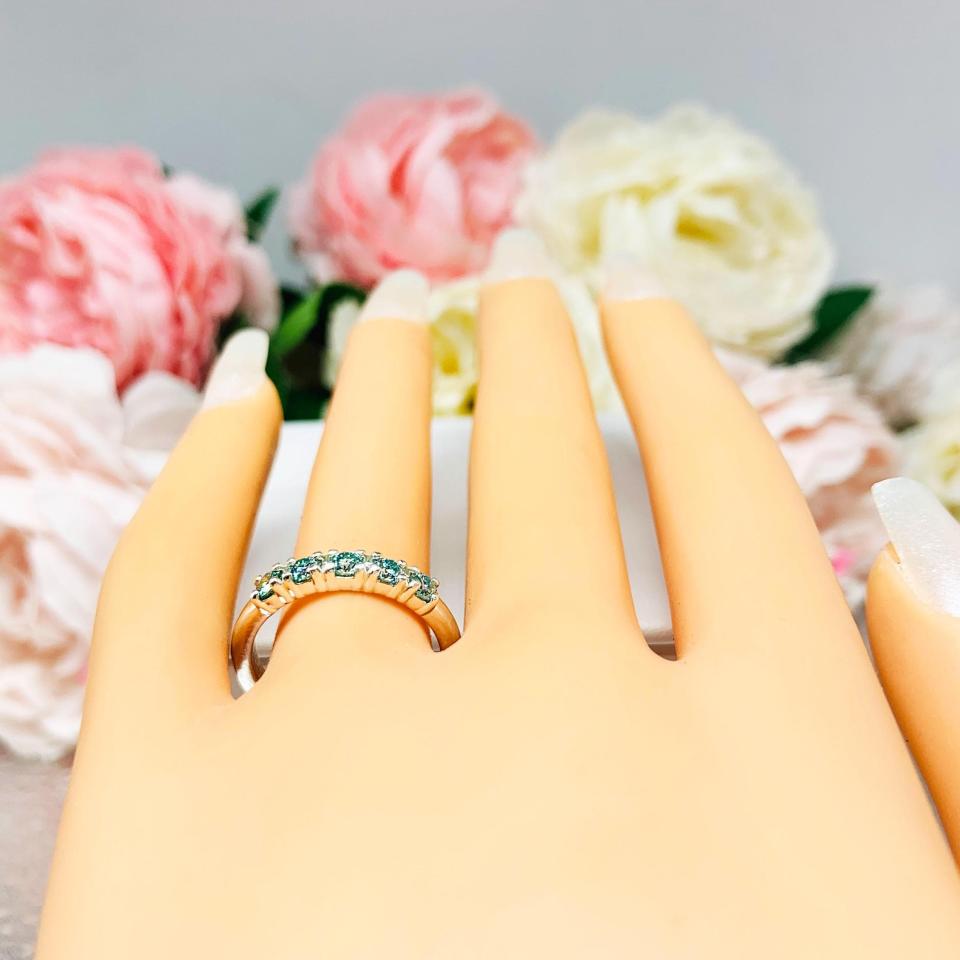 Mannequin hand with long white shimmery nails, displayed against a soft white-gray background with pink and white floral accents. The hand is wearing a sterling silver 5-stone band set with 3mm round sparkling aquamarine blue moissanites.