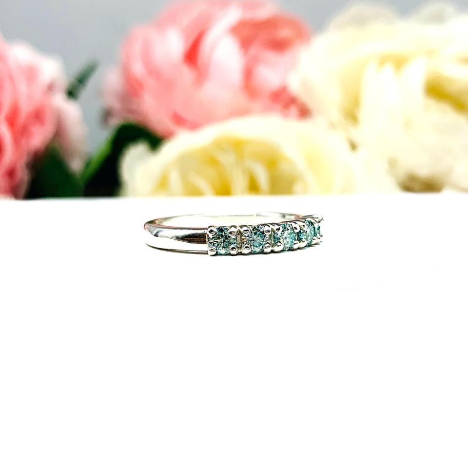 A sterling silver 5-stone band set with 3mm round sparkling aquamarine blue moissanites. Displayed on a white surface with a floral background.