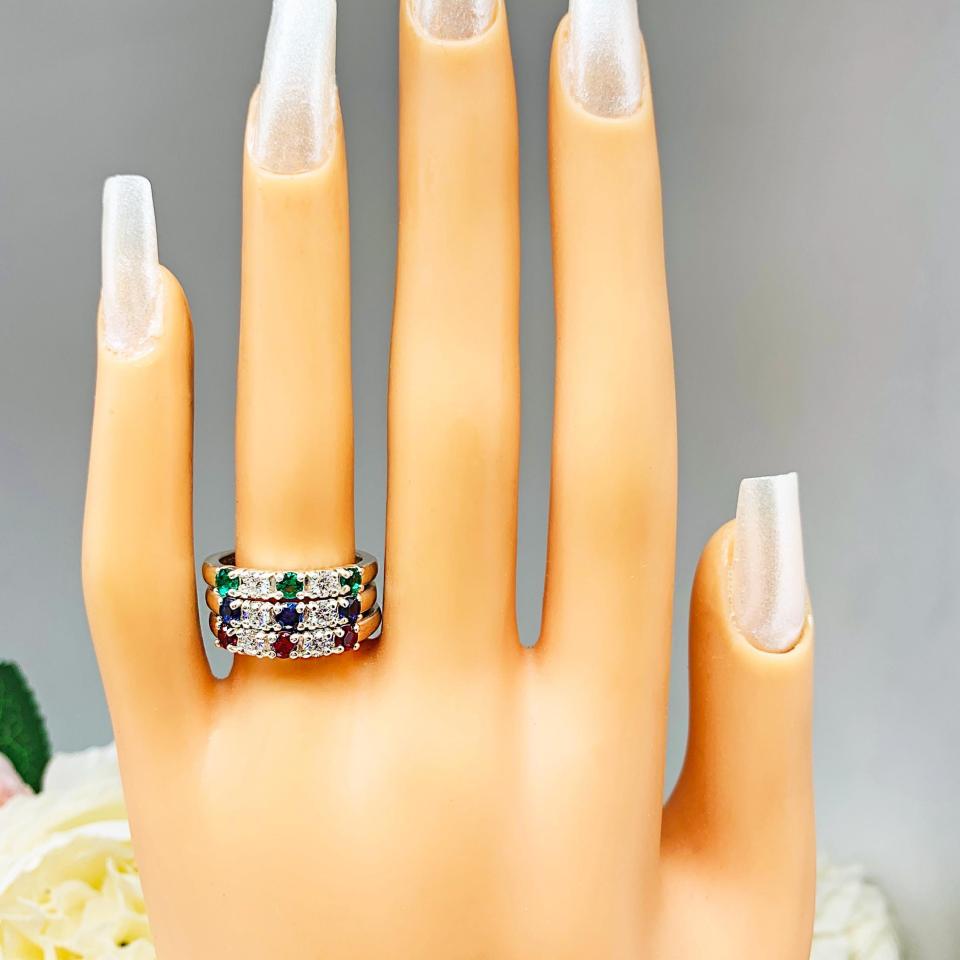 Three separate sterling silver bands displayed on a mannequin hand with long pearlescent nails. Each band features alternating 3mm round lab‑created gemstones: one with emerald and moissanite, one with blue sapphire and moissanite, and one with ruby and moissanite. Soft gray background with pink and white flowers.
