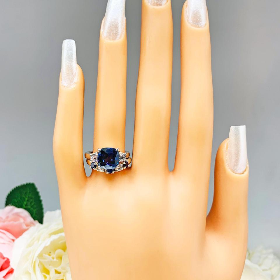 Mannequin hand with long white shimmery nails, displayed against a soft white-gray background with pink and white floral accents. The hand is wearing a sterling silver curved 5-stone band featuring alternating round lab blue sapphires and moissanites. The band has a classic design with the gemstones set in a row with three sapphires and two moissanites. It is featured here with a cushion-cut lab blue sapphire and cz ring (not included).