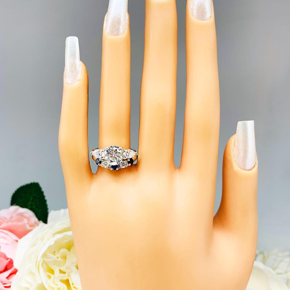 Mannequin hand with long white shimmery nails, displayed against a soft white-gray background with pink and white floral accents. The hand is wearing a sterling silver 5-stone band featuring alternating round lab blue sapphires and moissanites. The band has a classic design with the gemstones set in a row with three sapphires and two moissanites. It is featured here with a round-cut moissanite with pear sides three stone ring (not included).