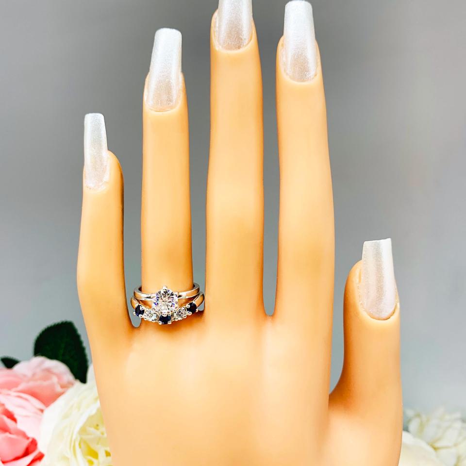 Mannequin hand with long white shimmery nails, displayed against a soft white-gray background with pink and white floral accents. The hand is wearing a sterling silver 5-stone band featuring alternating round lab blue sapphires and moissanites. The band has a classic design with the gemstones set in a row with three sapphires and two moissanites. It is featured here with a round-cut moissanite solitaire ring (not included).