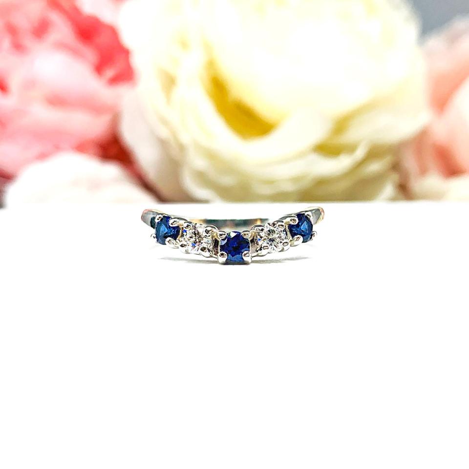 A sterling silver curved 5-stone band featuring alternating round lab blue sapphires and moissanites. The band has a classic design with the gemstones set in a row with three sapphires and two moissanites. Displayed on a white surface with a pastel floral background.