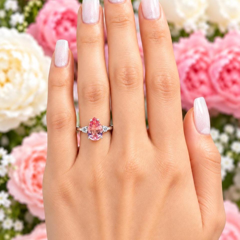 A woman's hand wearing a sterling silver ring with a 12x8mm pear lab-created padparadscha pink sapphire set in a 3-stone setting and flanked by a small colorless cubic zirconia on each side. Model image for size and styling reference — not a photo of the actual ring.