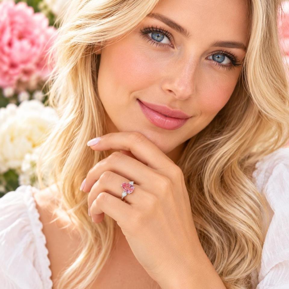 A woman wearing a sterling silver ring with a pear lab-created padparadscha pink sapphire set in a 3-stone setting and flanked by a small colorless cubic zirconia on each side. Model image for size and styling reference — not a photo of the actual ring.