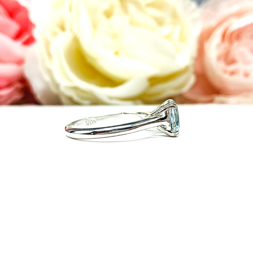 Sterling silver split-prong ring featuring a 7mm cushion-cut light blue aquamarine gemstone. Displayed against a soft-focus floral background, the ring sparkles with contrast and elegance.