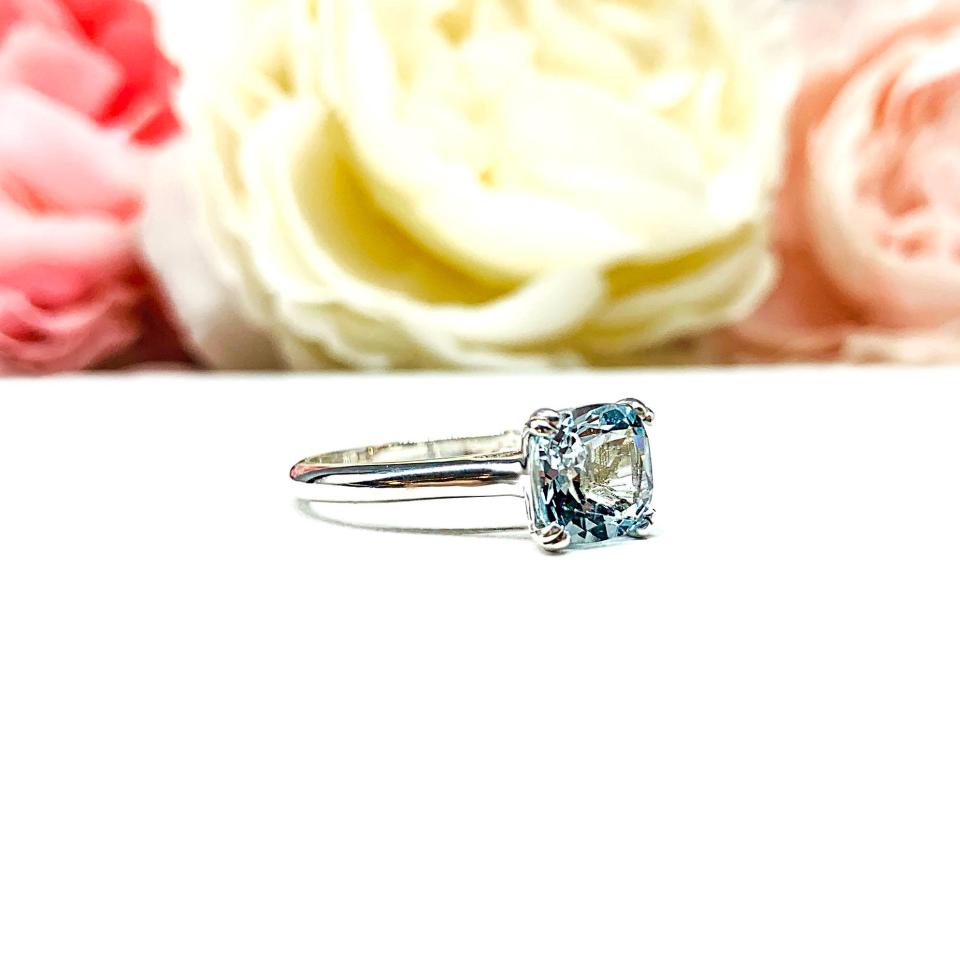 Sterling silver split-prong ring featuring a 7mm cushion-cut light blue aquamarine gemstone. Displayed against a soft-focus floral background, the ring sparkles with contrast and elegance.