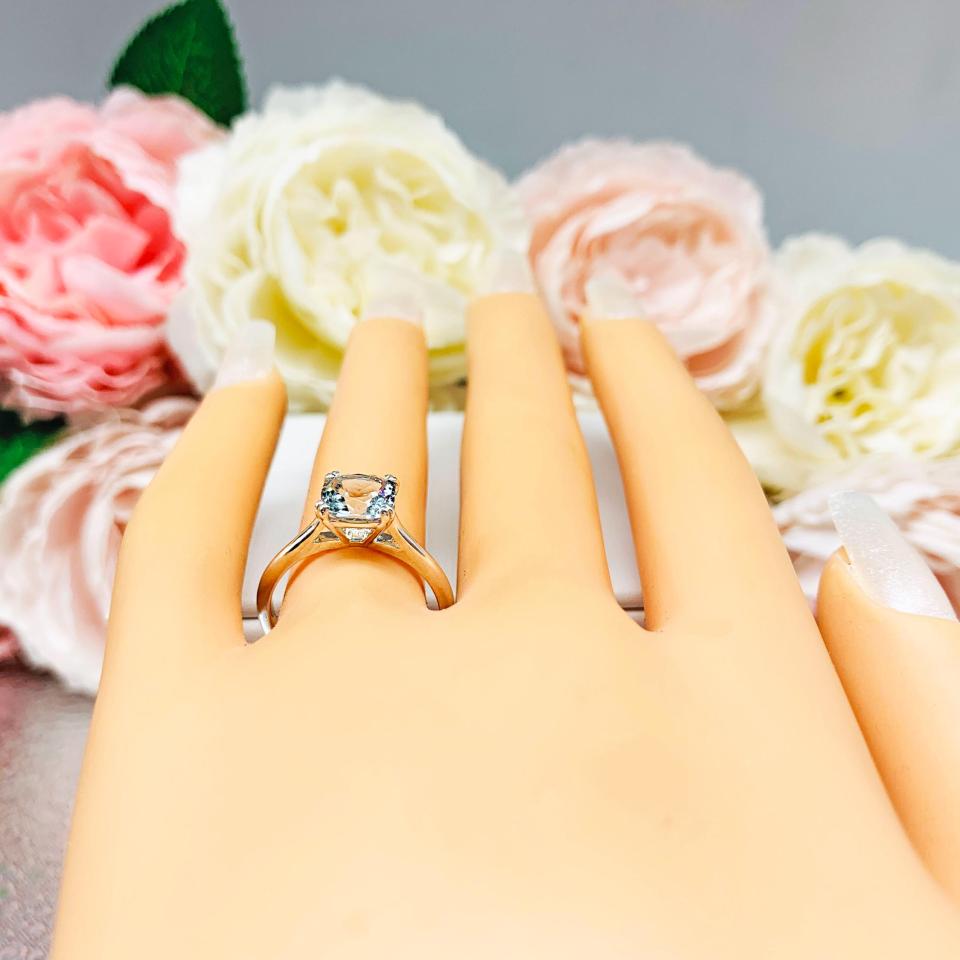 Mannequin hand with long white shimmery nails, displayed against a soft white-gray background with pink and white floral accents. The hand is wearing a sterling silver split-prong ring featuring a 7mm cushion-cut light blue aquamarine gemstone.