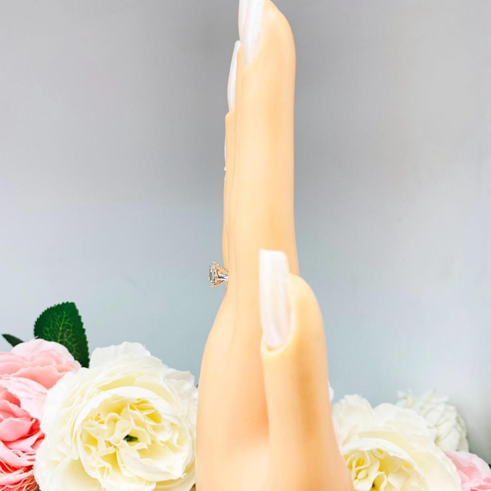 Mannequin hand with long white shimmery nails, displayed against a soft white-gray background with pink and white floral accents. The hand is wearing a sterling silver split-prong ring featuring a 7mm cushion-cut light blue aquamarine gemstone.