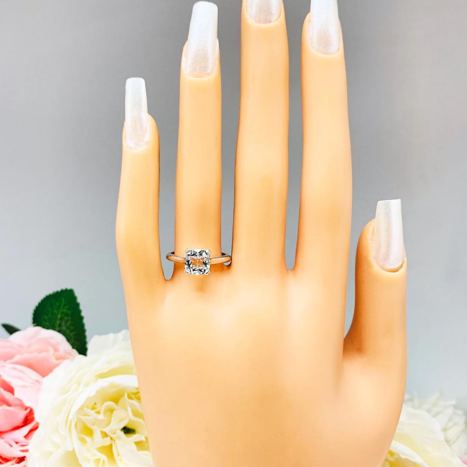 Mannequin hand with long white shimmery nails, displayed against a soft white-gray background with pink and white floral accents. The hand is wearing a sterling silver split-prong ring featuring a 7mm cushion-cut light blue aquamarine gemstone.