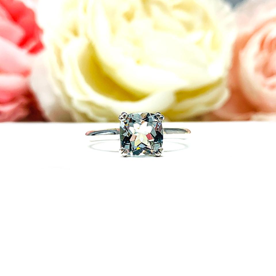Sterling silver split-prong ring featuring a 7mm cushion-cut light blue aquamarine gemstone. Displayed against a soft-focus floral background, the ring sparkles with contrast and elegance.