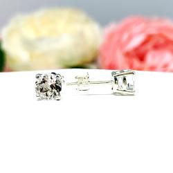 A pair of silver stud earrings with clear round white topaz gemstones. The earrings are on a white surface with a blurred background of pink and white flowers.