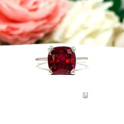 Sterling silver ring with a cushion cut lab created deep red ruby set in a split-prong setting set against a white surface with soft-focus pink and white flowers in the background.