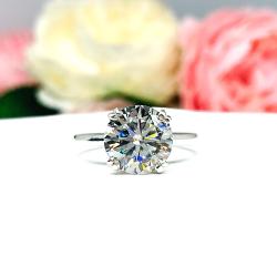 Sterling silver engagement ring featuring a brilliant 10mm (3.5 carat) round moissanite, shown in a close-up against a soft-focus background of pink and white flowers. The moissanite is set in a split prong cathedral setting.
