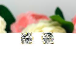 A pair of yellow gold stud earrings with brilliant cushion cut clear cubic zirconia stones. The basket style soft claw prong earrings are on a white surface