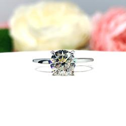 Close-up view of a near colorless (G/H color) round brilliant cut cubic zirconia solitaire ring in a claw prong cathedral setting. The ring is made of sterling silver and features a sleek, minimalist design. The setting is simple yet elegant, allowing the center stone to take center stage. The ring is displayed against a soft, blurred background of pastel-colored flowers, creating a romantic and delicate ambiance.