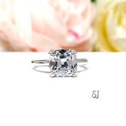 A sterling silver split-prong cathedral engagement ring set with a colorless old mine cut cubic zirconia. The ring is set against a white background with a blurred image of pink and white flowers in the background.