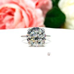 Close-up view of a near colorless (G/H/I color) round brilliant cut cubic zirconia solitaire ring in a split prong cathedral setting. The ring is made of sterling silver and features a sleek, minimalist design. The setting is simple yet elegant, allowing the center stone to take center stage. The ring is displayed against a soft, blurred background of pastel-colored flowers, creating a romantic and delicate ambiance.
