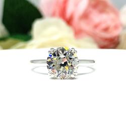 A romantic close-up of a 9mm (3 ct) old European cut near-colorless CZ solitaire engagement ring. Set in a sterling silver split prong cathedral-style mount, the ring gleams with vintage-inspired elegance. Soft pink and white flowers blur in the background, adding a dreamy, bridal ambiance.