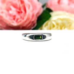 Sterling silver band featuring a 5×3 mm emerald‑cut green tourmaline set flush in a polished band, displayed on a white surface with soft pink and cream flowers blurred in the background.