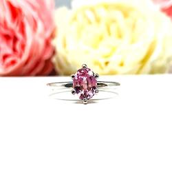 Sterling silver ring with an 8x6mm oval lab created padparadscha sapphire set in a 6-prong setting set against a white surface with soft-focus pink and white flowers in the background.