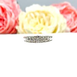 A sterling silver graduated moissanite engagement ring featuring a 4mm center stone flanked by 3mm and 2.5mm side stones, shown in a close-up against a soft-focus background of pink and white flowers. The moissanites sparkle with icy brilliance, making this an elegant alternative engagement or anniversary ring.