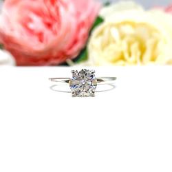 A solitaire engagement ring with a round brilliant DEF moissanite. The setting is a classic four-prong design, and the band is a simple, sterling silver band. The ring is displayed on a white background with blurred pink and yellow flowers in the background.