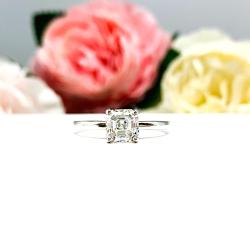 Sterling silver 4-prong asscher cut DEF moissanite solitaire engagement ring. The ring is resting on a white surface with a background of pink and white flowers.