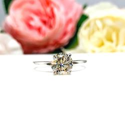 A solitaire engagement ring with a round brilliant light champagne moissanite. The setting is a classic four-prong design, and the band is a simple, sterling silver band. The ring is displayed on a white background with blurred pink and yellow flowers in the background.