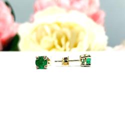 A pair of 14k yellow gold stud earrings set with 4mm round natural emerald gemstones. The four prong basket earrings are on a white surface with the left earring facing front to showcase the clarity and vibrant green color of the stones, and the right earring facing toward the right to display the center post and butterfly friction clasp.