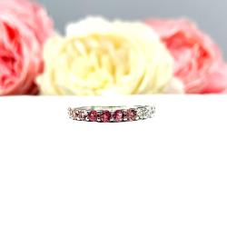 Sterling silver twelve stone band set with 2.5mm round gemstones in this configuration: four white topaz, four pink tourmaline and four light pink tourmaline set in a shared prong setting, displayed on a white surface with soft-focus pastel flowers in the background.