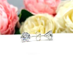 A pair of sterling silver 3-prong martini stud earrings with 6.5mm colorless old European cut cubic zirconias. The earrings are resting on a white surface with a background of pink and white flowers.