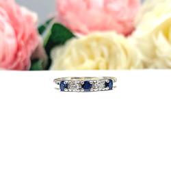 A sterling silver 5-stone band featuring alternating round lab blue sapphires and moissanites. The band has a classic design with the gemstones set in a row with three sapphires and two moissanites. Displayed on a white surface with a pastel floral background.