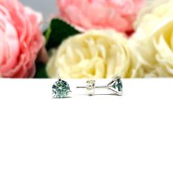 Sterling silver stud earrings featuring round authentic sea foam green moissanite stones in martini-style settings, displayed on a white surface with soft floral elements in the background.